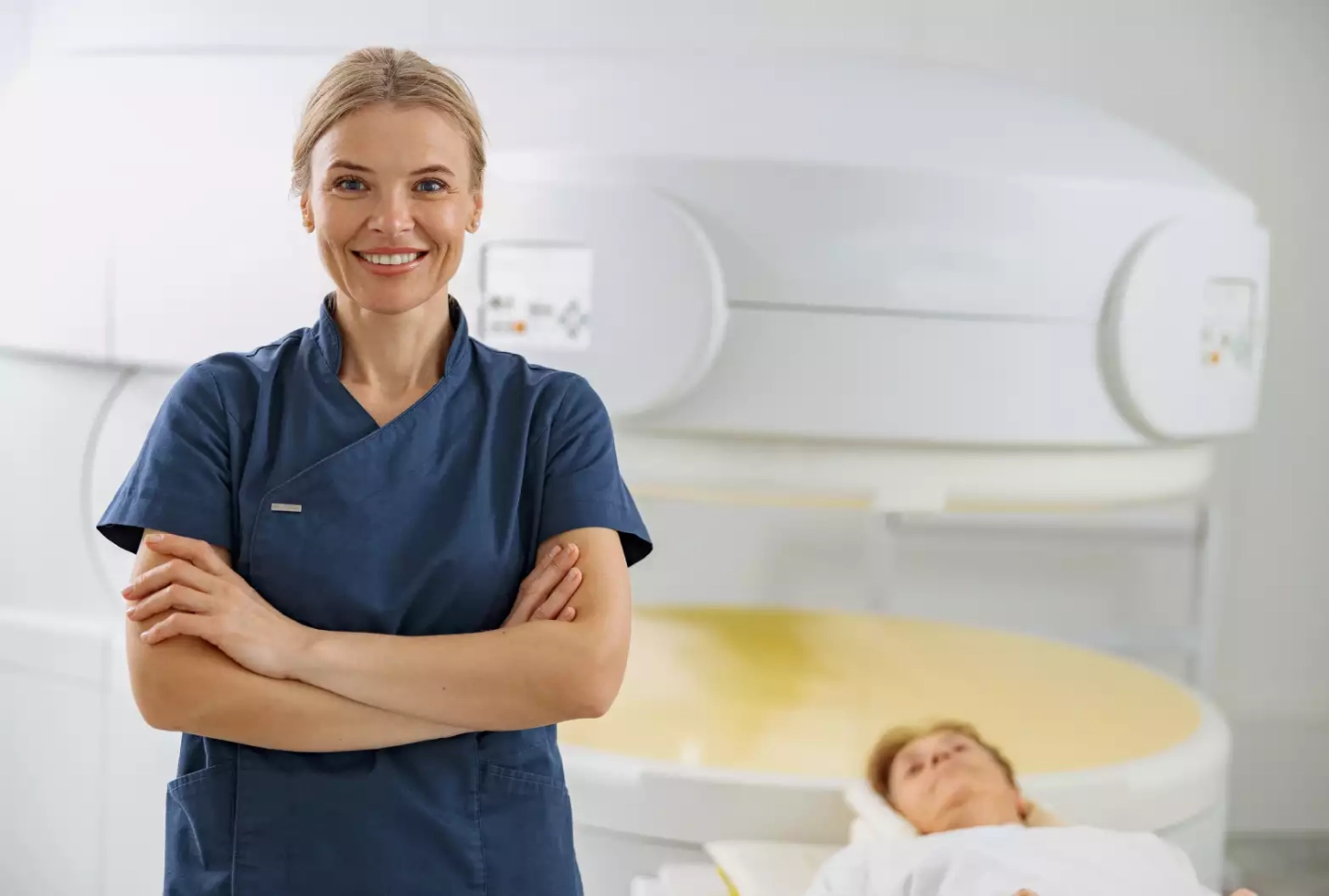 Medizinische Fachangestellte lächelt in Kamera, im Hintergrund liegt eine Patientin vor dem MRT
