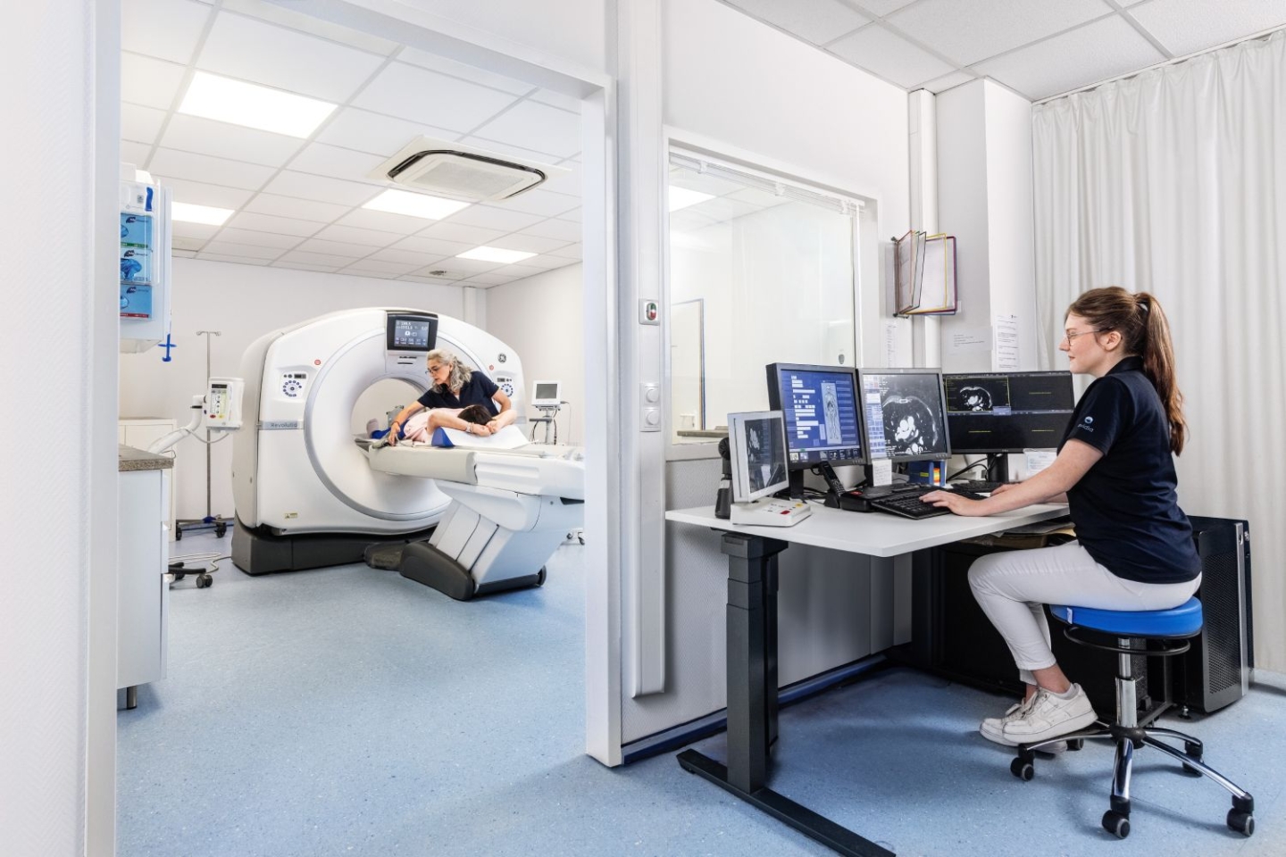 Dieses Bild zeigt eine medizinische Untersuchungsszene in einem CT-Raum. Auf der linken Seite des Bildes liegt ein Patient auf einer Liege und wird in die Röhre eines  MRT-Geräts geschoben, während eine medizinische Fachkraft dabei hilft. Auf der rechten Seite des Bildes sitzt eine medizinische Fachkraft an einem Schreibtisch mit mehreren Bildschirmen, auf denen MRT-Bilder zu sehen sind. Die Fachkraft überwacht und steuert die Untersuchung. Der Raum ist sauber und modern eingerichtet.