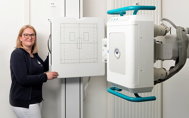 Eine lächelnde MTRA steht neben einem Röntgengerät in einem Untersuchungsraum.