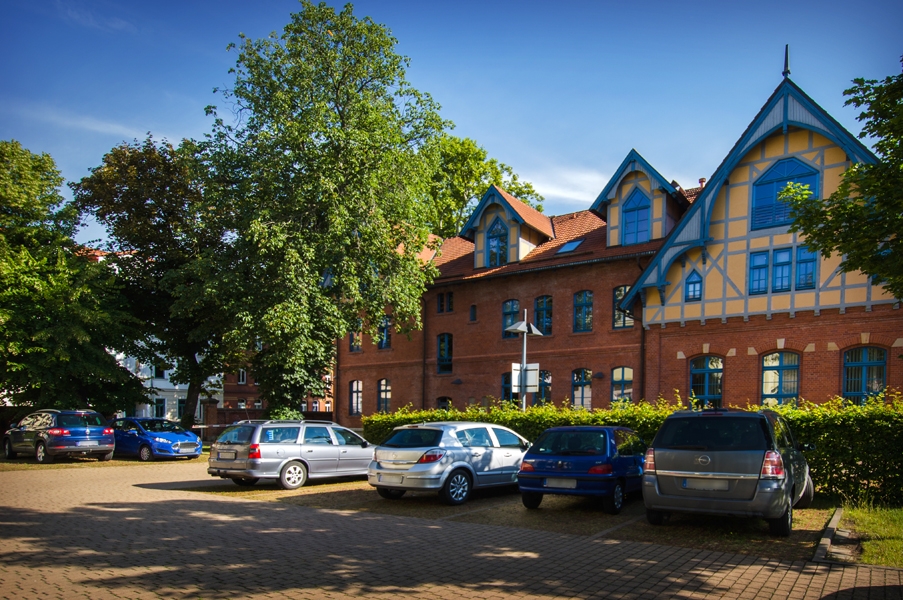 Außenaufnahme eines Backsteingebäudes an einem sonnigen Tag. Vor dem Gebäude befindet sich ein Parkplatz mit mehreren Autos. Links und rechts stehen hohe, grüne Bäume.