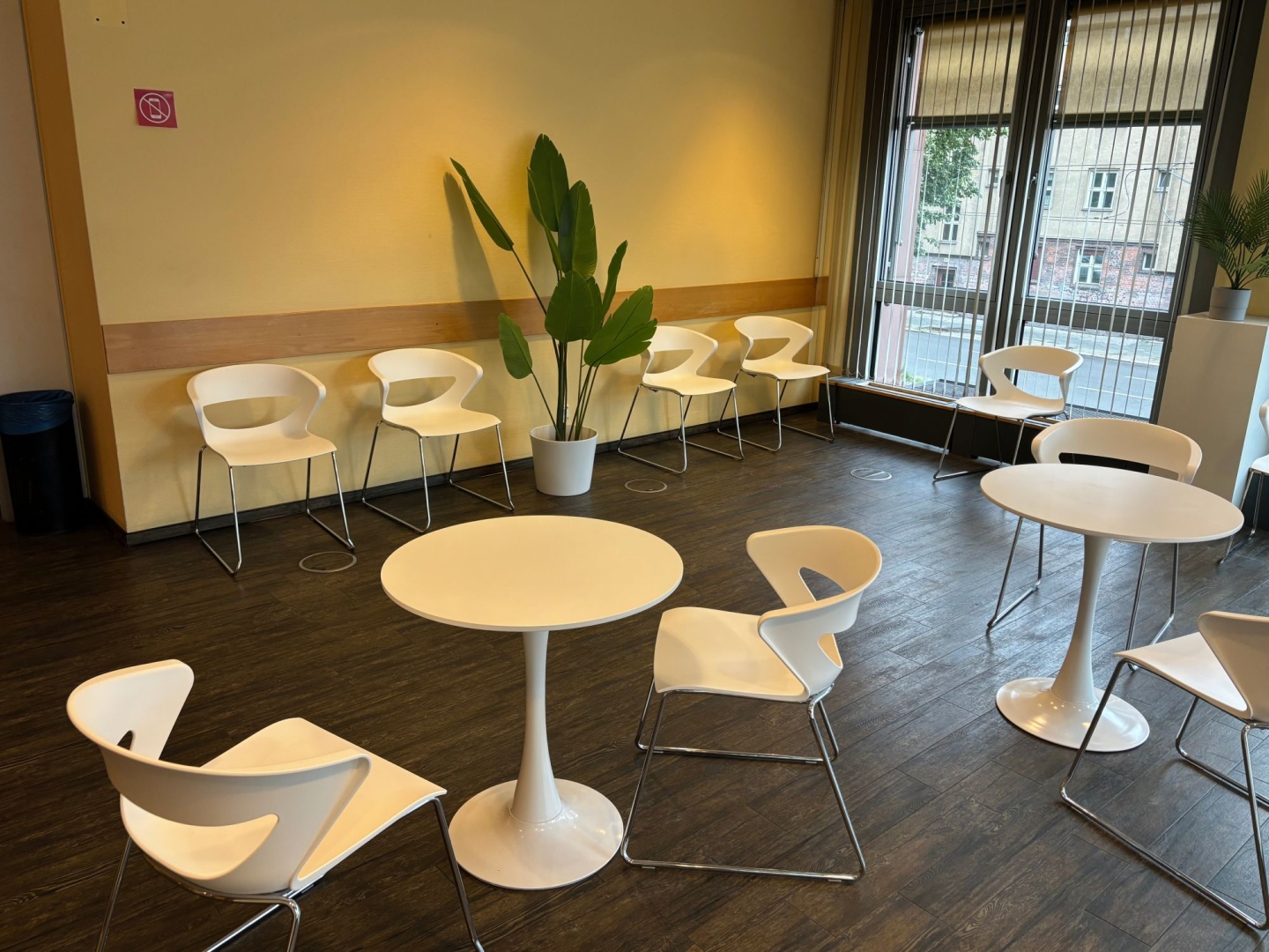 Innenraum Wartebereich, mit dutzenden modernen weiße Stühlen und einigen runden Tischen. Dunkler Holzboden und eine Fensterfront.