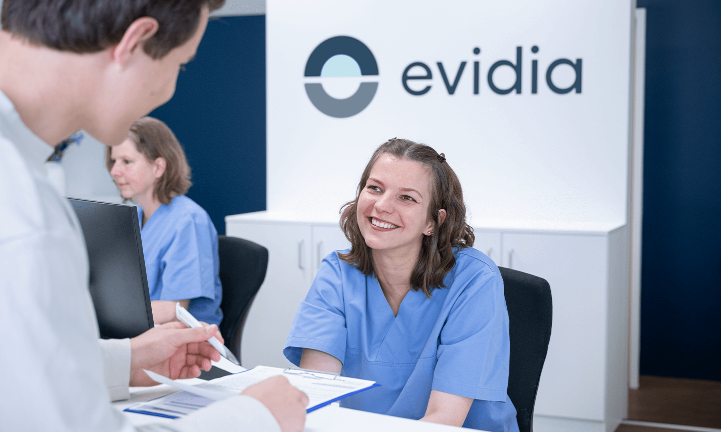 Mitarbeiterin des MVZ lächelt Person, die etwas an einem Klemmbrett ausfüllt, an.