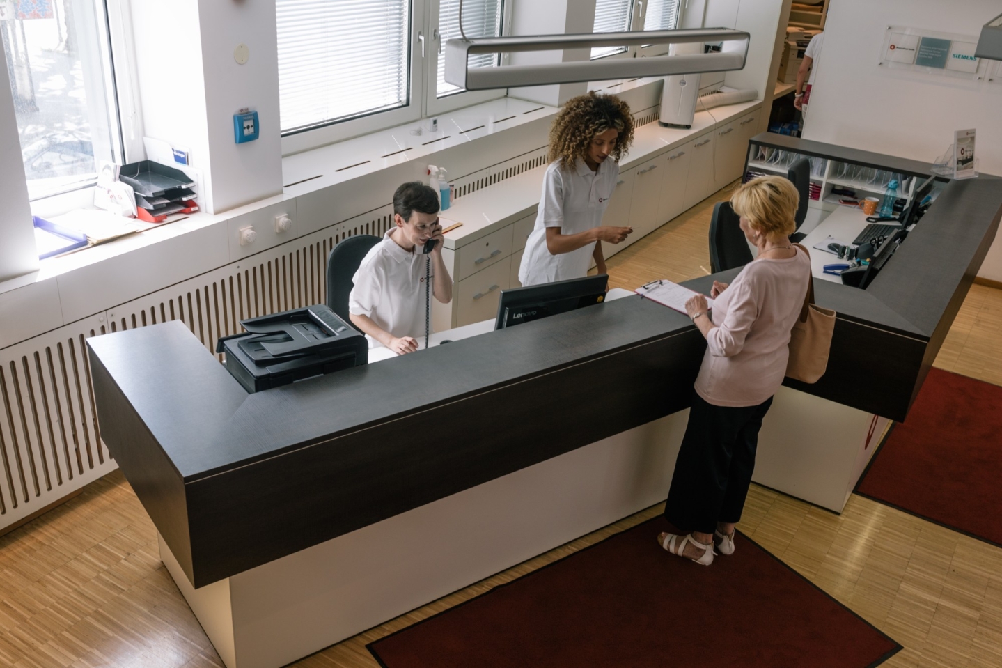 Empfangstresen einer Praxis. Zwei Mitarbeitende hinter dem Tresen, sowie eine Patientin interargieren.