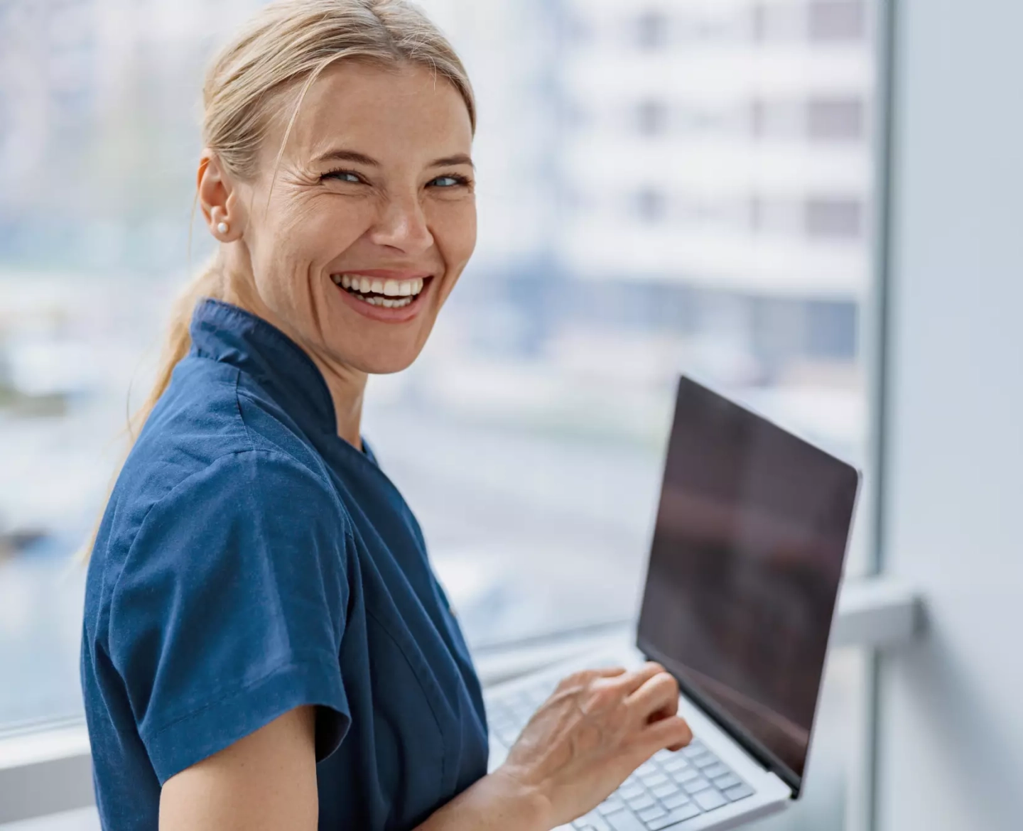 Frau in blauem Shirt und Laptop in der Hand lacht in die Kamera