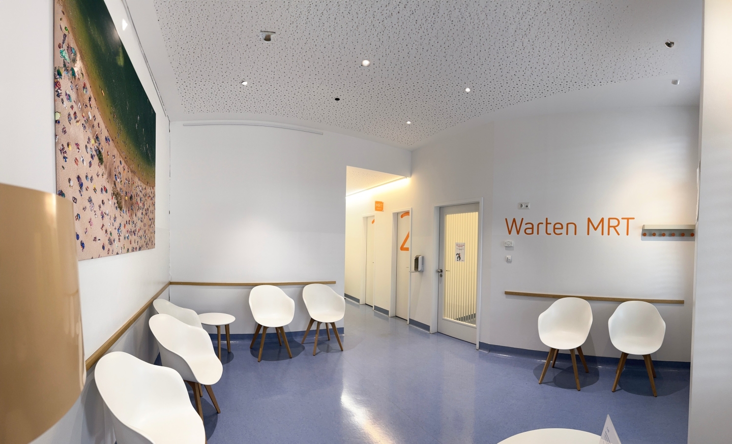 Helles Wartezimmer mit blauen Boden. An der Wand hängt ein großes Bild einer Strandszene, an der anderen steht 'Warten MRT'. Es stehen weiße Stühle an den Wänden.