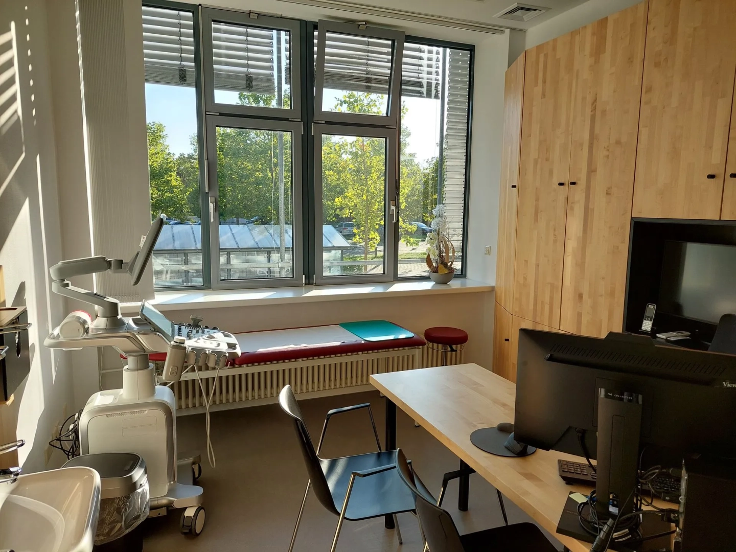 Ein Sprechzimmer mit großer Fensterfront, einer Liege, einem Schreibtisch mit PC. Viel Licht fällt durch die Fenster in den Raum.