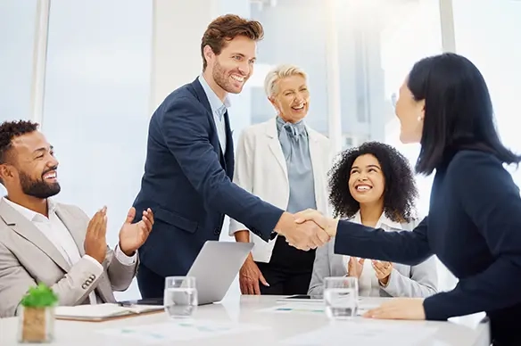 Zwei Personen geben sich im Business-Kontext die Hand, weitere Personen schauen zu und applaudieren