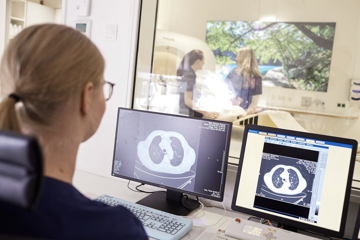 Eine Mitarbeitende schaut auf zwei Monitoren auf verschiedene Scans. Zwei weitere Mitarbeitende stehen hinter einer Glaswand und helfen dem Patienten.