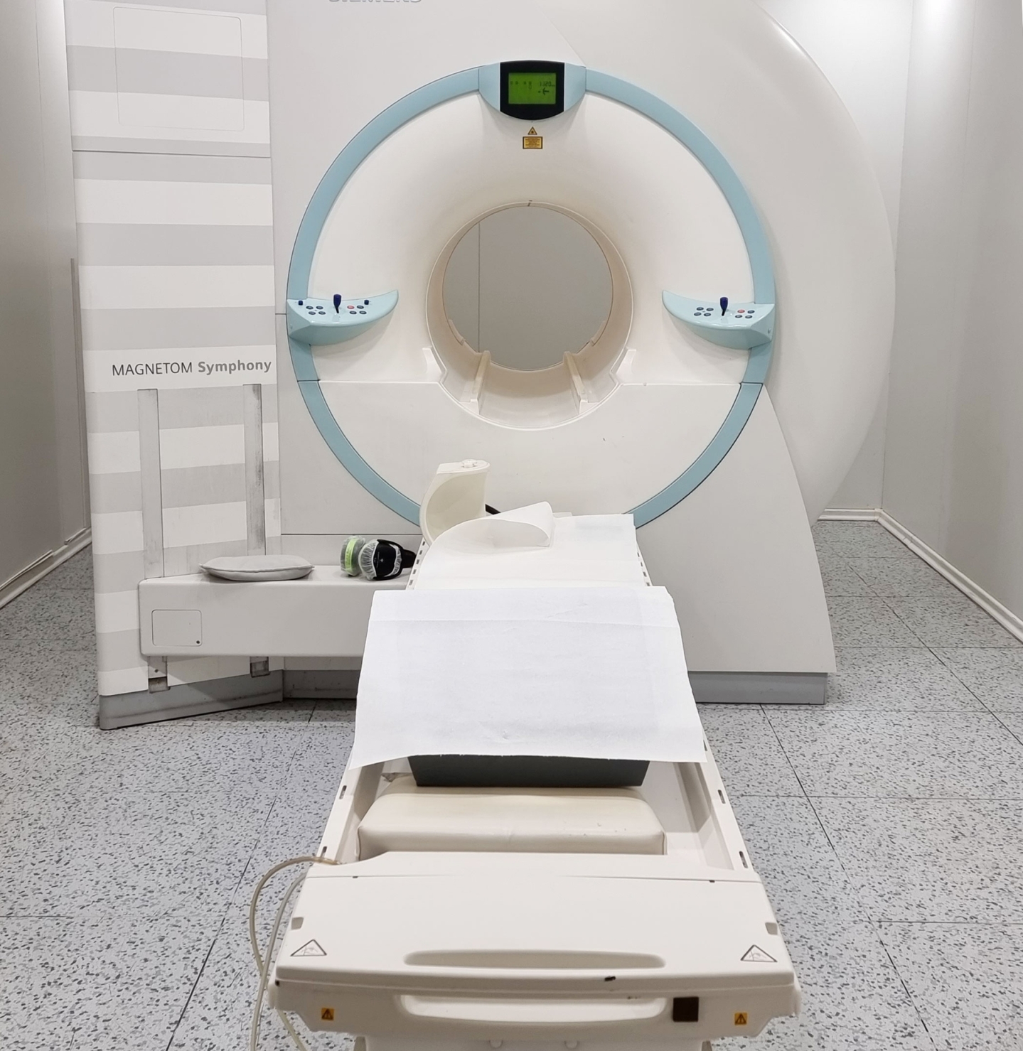 Ein MRT Gerät in einem Untersuchungsraum. Der Raum hat helle Wände und eine grauen, gefliesten Boden.