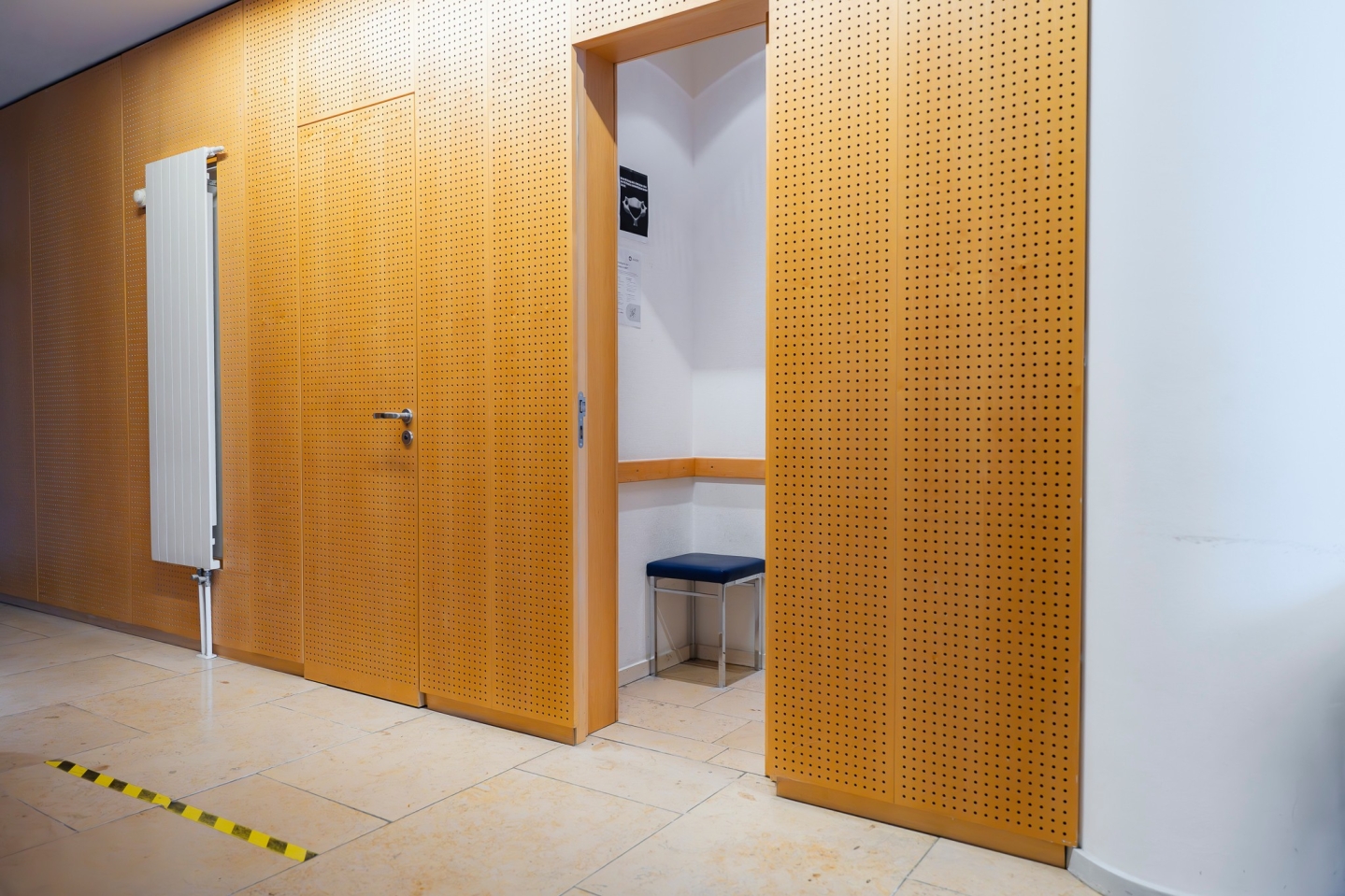 Heller Flur mit einer gelochten Holzfaserwand. Ein offener Durchgang gibt den Blick frei auf eine kleine Kabine mit einem blau gepolsterten Hocker. Links an der Wand hängt ein weißer Heizkörper, auf dem beigen Fliesenboden klebt ein gelb-schwarzer Markierungsstreifen.