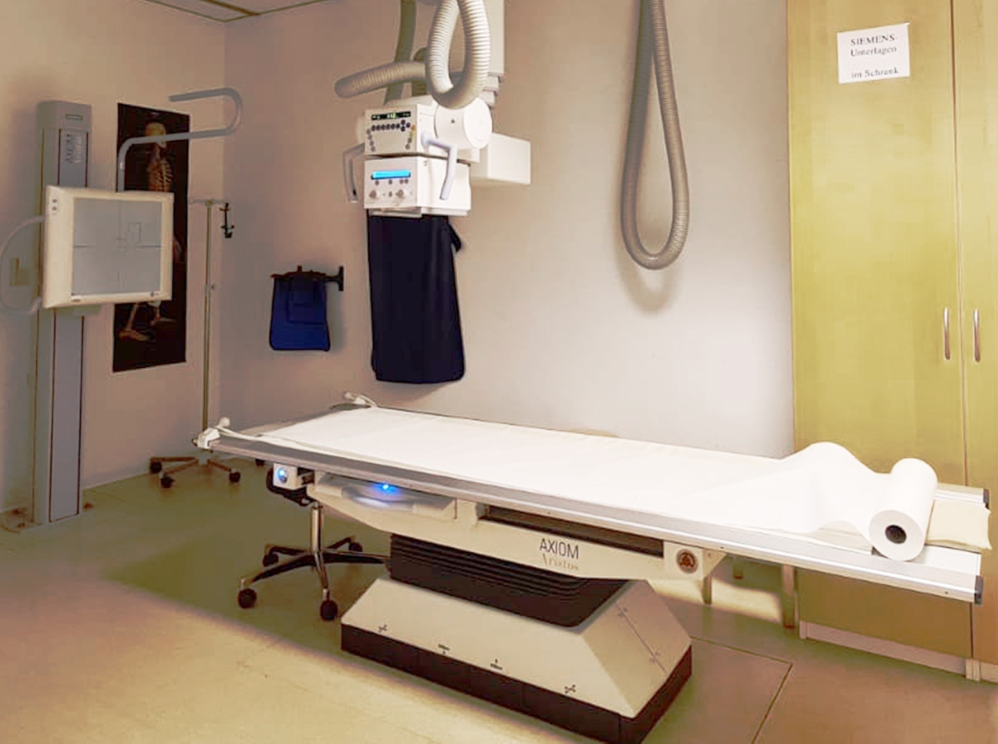 Ein Röntgenuntersuchungszimmer mit einer zentralen, weißen Patientenliege, darüber eine Röntgenröhre.