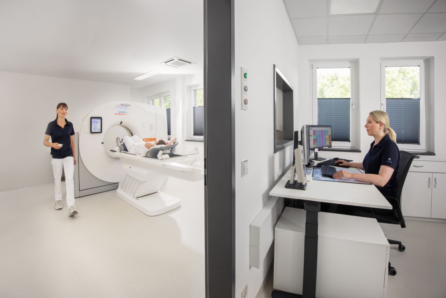 Dieses Bild zeigt eine medizinische Untersuchungsszene in einem CT-Raum. Auf der linken Seite des Bildes liegt ein Patient auf einer Liege, die in die Röhre eines Siemens Magnetom Prisma CT-Geräts geschoben wird. Eine medizinische Fachkraft steht daneben und bereitet die Untersuchung vor. Auf der rechten Seite des Bildes sitzt eine andere medizinische Fachkraft in einem separaten Raum an einem Schreibtisch mit mehreren Monitoren, auf denen CT-Bilder zu sehen sind. Sie überwacht und steuert die Untersuchung von dort aus. Der Raum ist hell und modern eingerichtet, mit weißen Wänden und Fußböden, sowie Fenstern, die natürliches Licht hereinlassen.