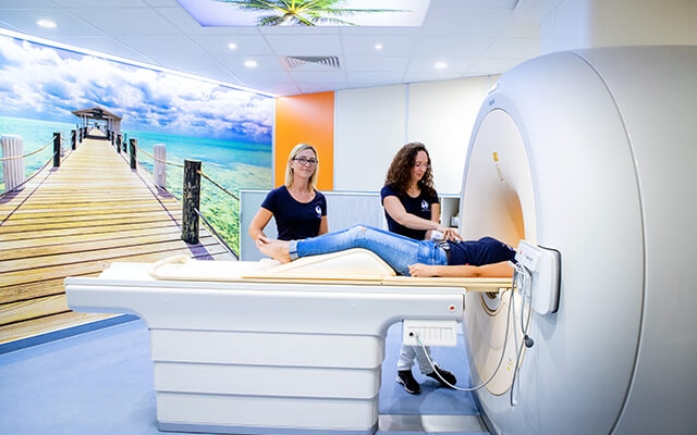 Zwei Mitarbeitende positionieren einen Patienten auf der Liege eines MRT-Gerätes. Im Hintergrund ist eine Wandtapete mit einer tropischen Szene.