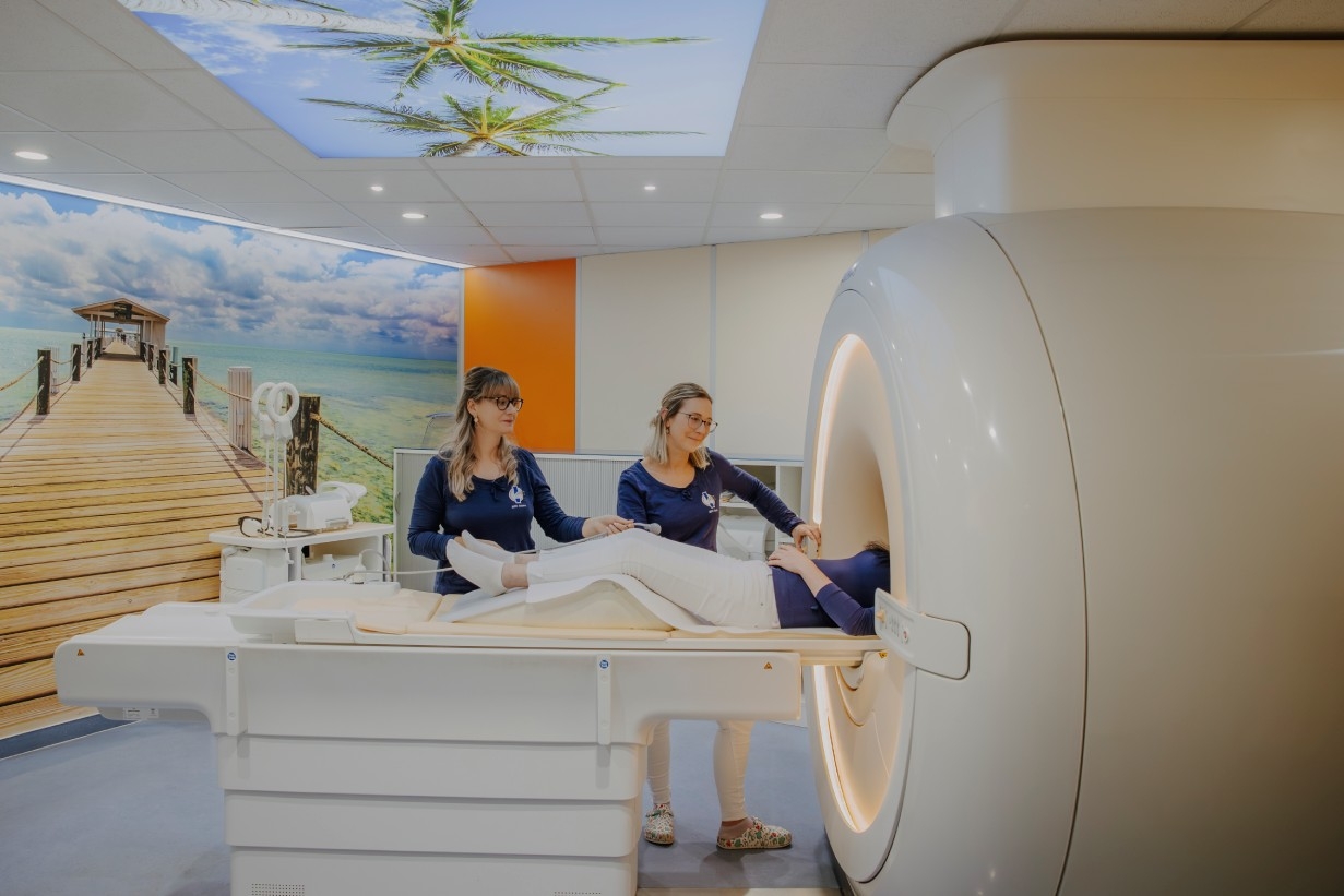 Zwei Mitarbeitende positionieren einen Patienten auf der Liege eines MRT-Gerätes. Im Hintergrund ist eine Wandtapete mit einer tropischen Szene.