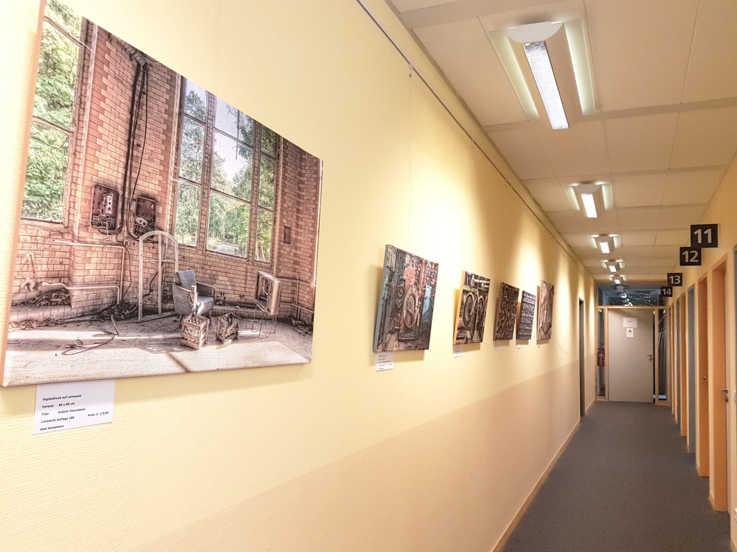 Ein heller Flurin einer Praxis. An der linken Wand hängen mehrere Bilder, rechts sind benummerte Türen.