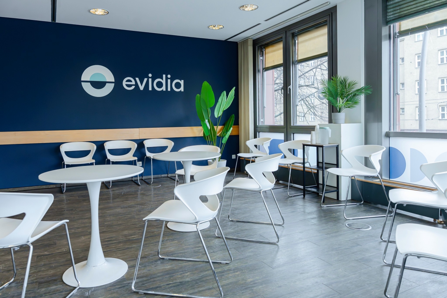 Ein großzügiger, moderner Wartebereich der Marke „evidia“ mit dunklem Bodenbelag. Eine auffällige, dunkelblau gestrichene Wand trägt das Firmenlogo in Weiß. Im Raum sind zahlreiche futuristisch geformte, weiße Stühle mit Metallbeinen um zwei runde weiße Tische herum angeordnet. Große Fenster sorgen für Tageslicht, und Zimmerpflanzen setzen grüne Akzente.