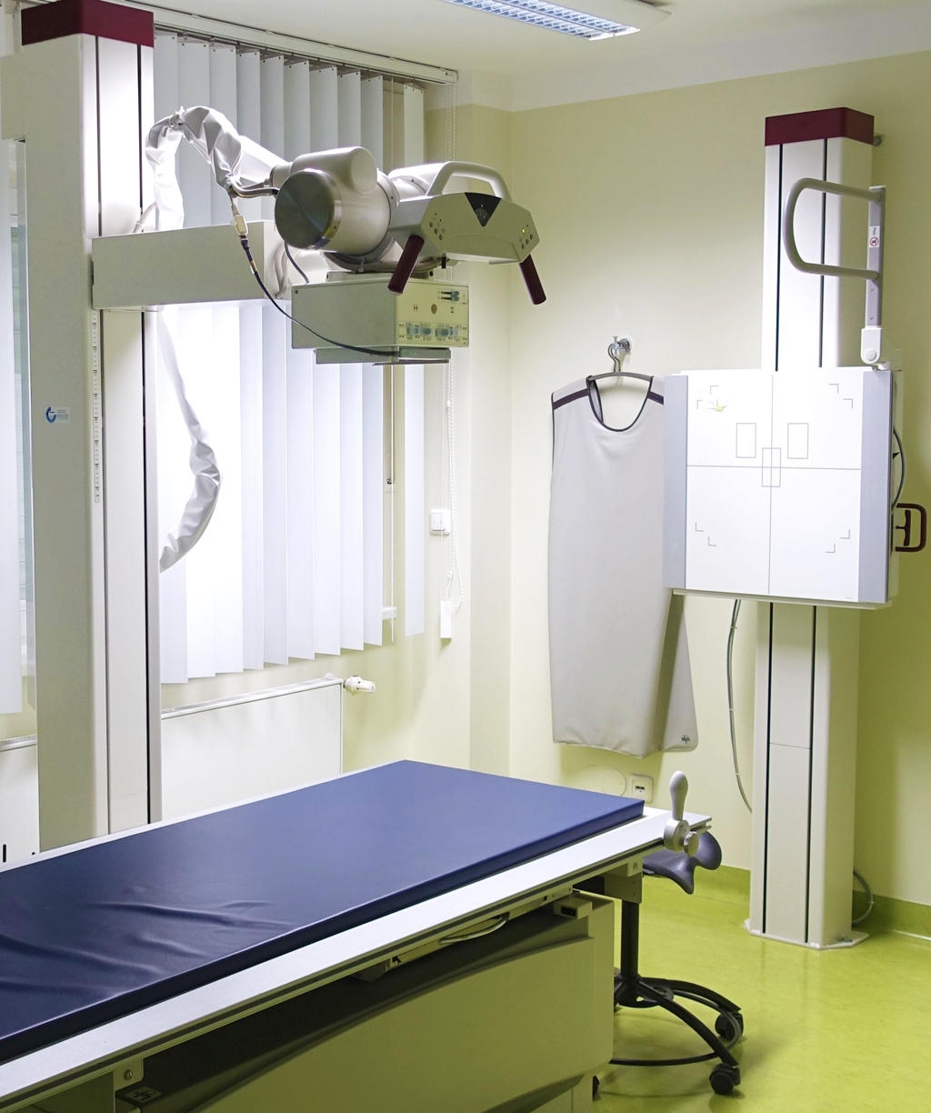 Untersuchungsraum für radiologische Verfahren mit einem grünen Boden. Im Raum befindet sich ein blau bezogener Untersuchungstisch und eine Röntgenröhre.