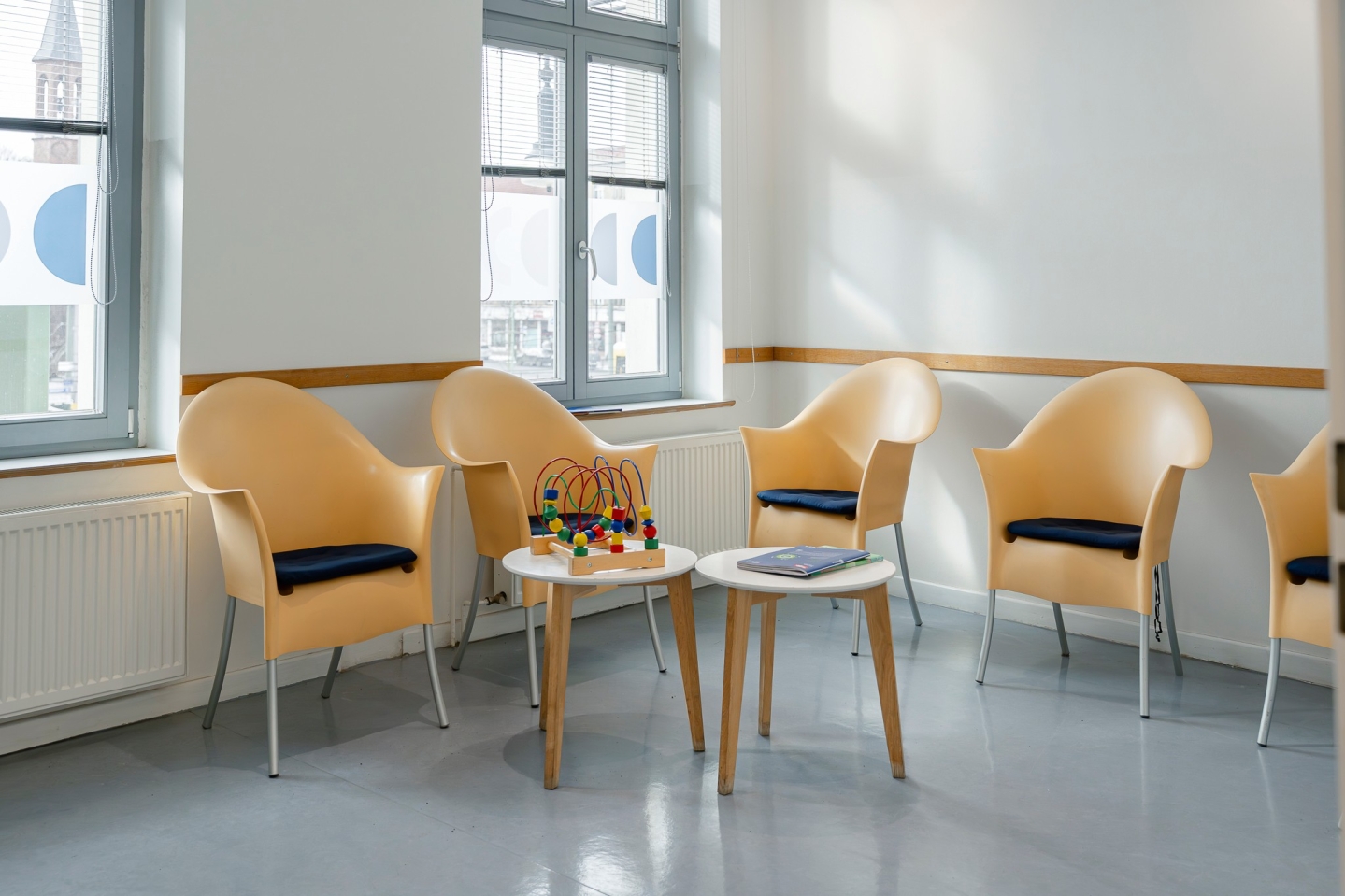 Ein heller Wartebereich mit grauem Boden und mehreren beige-gelben Schalensesseln, die mit dunklen Sitzkissen ausgestattet sind. In der Mitte stehen zwei kleine, runde Holztische – einer davon mit einer bunten Motorikschleife für Kinder. Im Hintergrund befinden sich große Fenster mit Jalousien und blau-weißem Sichtschutz.