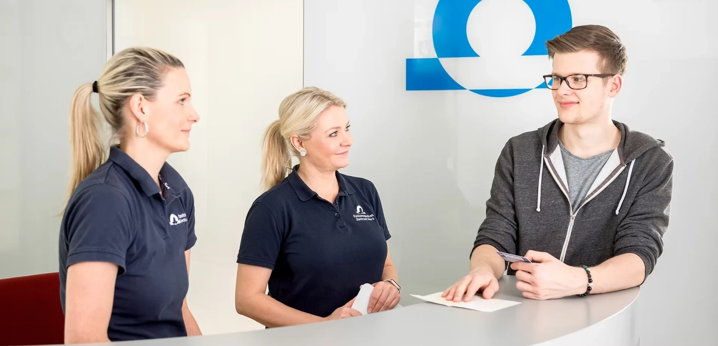 Zwei Fachkräfte stehen am Tresen mit einem Patienten.