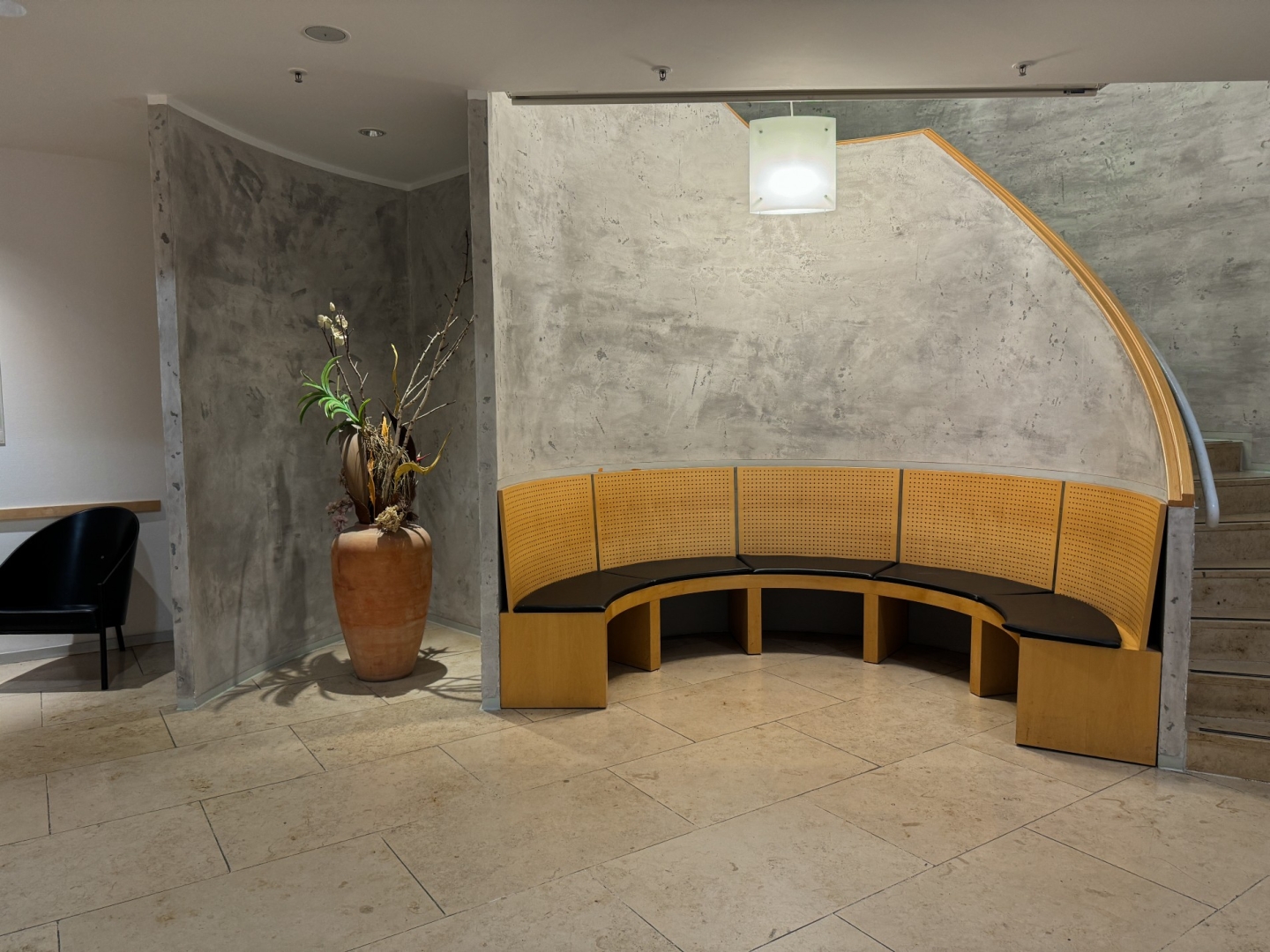 Eine Sitznische im Wartebereich der Praxis. Eine halbkreisförmige Bank mit schwarzen Polstern. Die Wände sind in Betonoptik, hinter der Sitzbank führt eine Treppe nach oben.