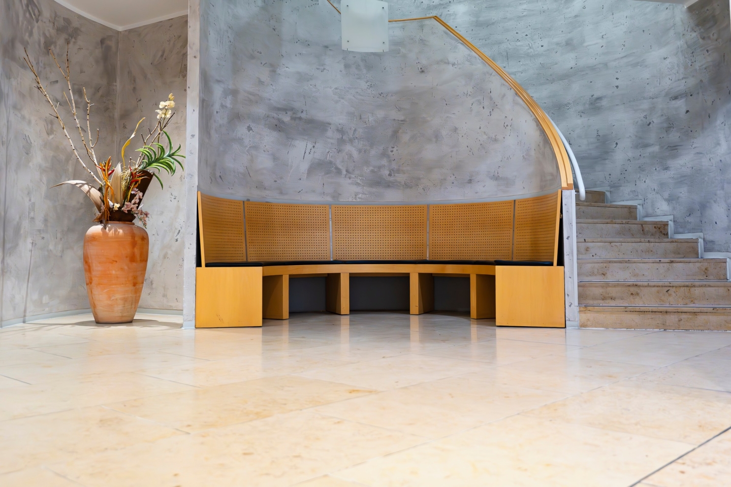 Ein moderner Eingangsbereich oder Foyer mit beigen Bodenfliesen und Wänden in grauer Betonoptik. Im Zentrum steht eine geschwungene, hölzerne Sitzbank mit gelochter Rückenlehne. Rechts führt eine Treppe nach oben, links sorgt eine große Terrakotta-Vase mit Blumengesteck für Dekoration.