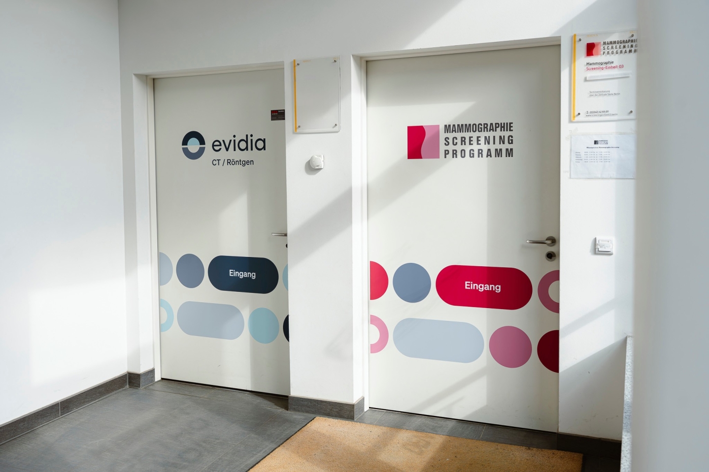 Zwei nebeneinanderliegende, weiße Praxistüren mit Beschriftung. Die linke Tür zeigt das Logo „evidia CT / Röntgen“ mit blau-grauen Mustern und einem dunkelblauen „Eingang“-Schild. Die rechte Tür ist als Zugang zum „Mammographie Screening Programm“ mit pink-grauen Mustern und einem pinken „Eingang“-Schild gekennzeichnet.