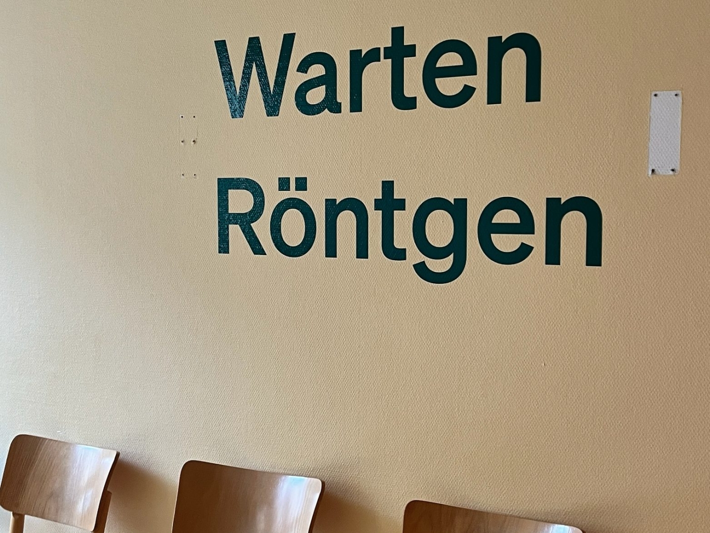 Wartebereich. An der Wand steht Warten Röntgen, darunter mehrere Holzstühle.