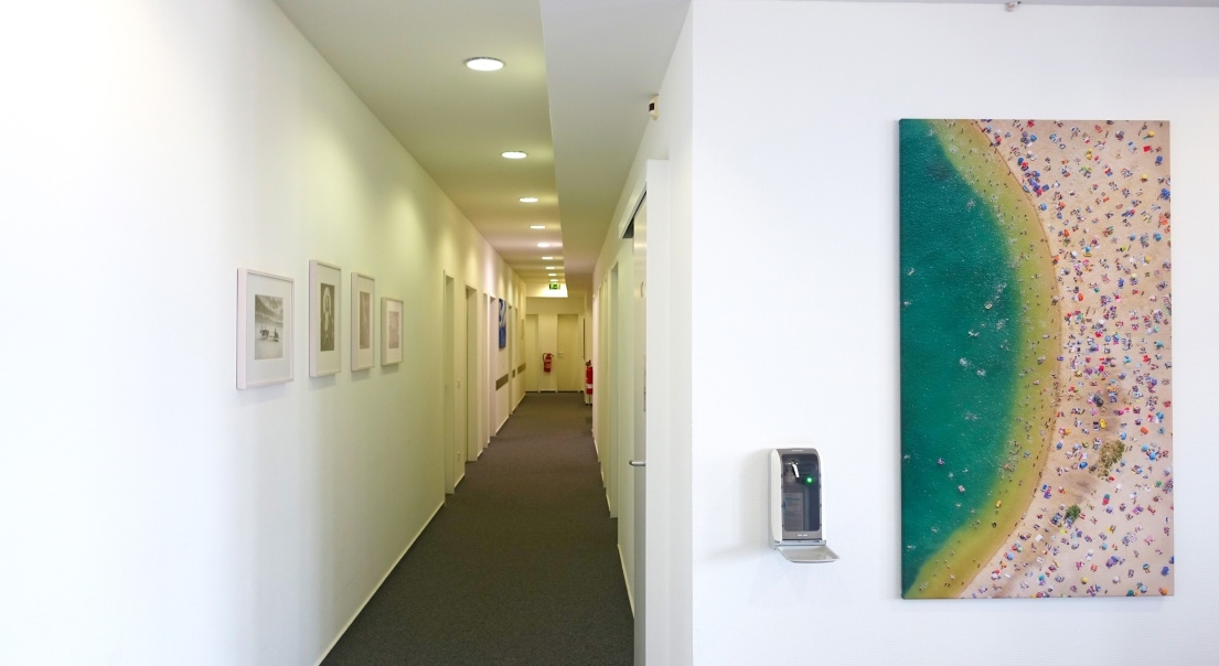 Langer, heller Flur in einer Praxis. An der rechten Wand hängt ein großes Gemälde, links sind mehrere, kleinere, gerahmte Bilder in einer Reihe angeordnet.
