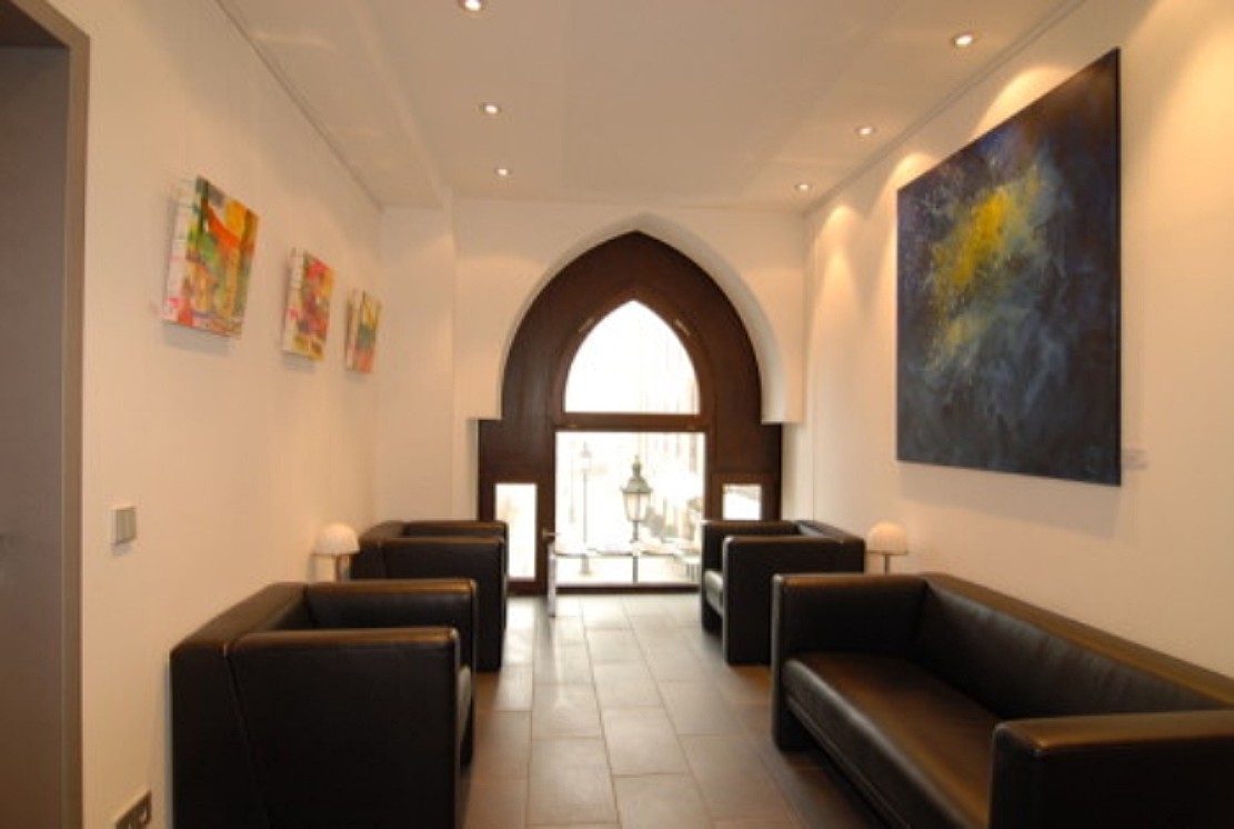 Heller Wartebereich durch ein großes Fenster mit bequemen Sesseln und einer Couch. Gemälde an den Wänden.