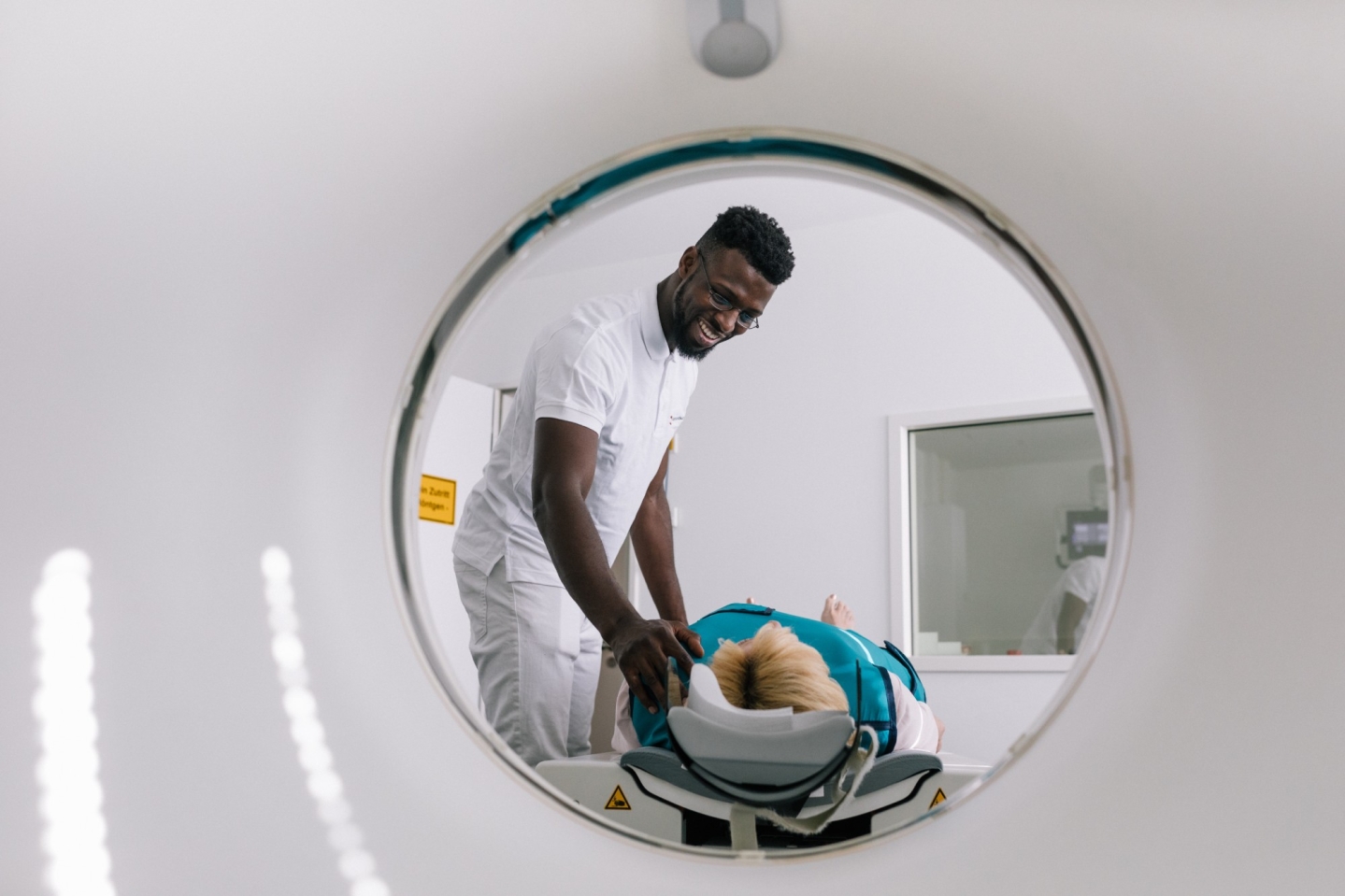Szene gezeigt durch die Runde Öffnung eines medizinisches Gerätes. Ein lächelnder Mann in weißer medizinischer Kleidung positioniert eine liegende Patientin.