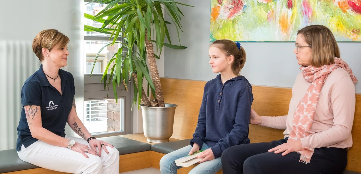 Mitarbeiterin sitzt im Wartebereich und redet mit junger Patientin und Mutter.