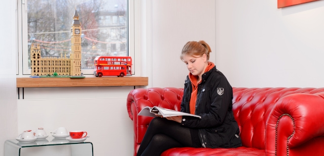 Eine Frau sitzt auf einem roten Sofa im Wartebereich der Praxis und liest eine Zeitung.